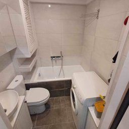 Roman Graczyk - Wykończona łazienka w jasnych płytkach z wanną, umywalką, toaletą i pralką, widok z perspektywy drzwi, z akcentem na minimalistyczne szafki i armaturę.