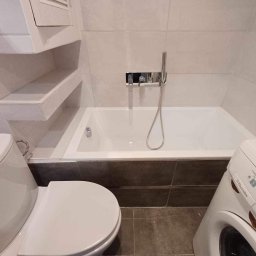 Roman Graczyk - Wykończona łazienka z białą wanną, toaletą, zabudowanymi półkami i pralką Electrolux, płytki w odcieniach bieli i szarości.