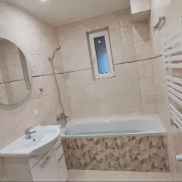 Wykończona łazienka w beżowych płytkach z dekoracyjnym pasem na wannie, okrągłe podświetlane lustro, biała umywalka z szafką i grzejnik drabinkowy.