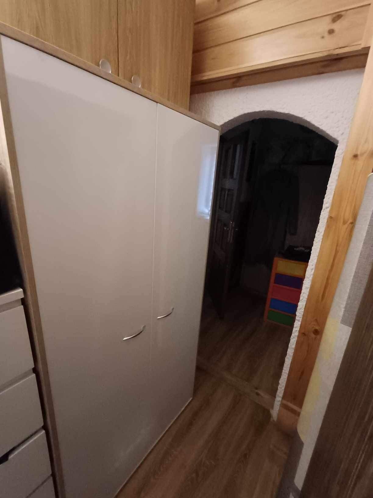 Biała szafa z połyskiem i drewnianym obramowaniem, umieszczona w pomieszczeniu z drewnianą boazerią i podłogą z paneli, widok na ciemne przejście z kolorową komodą.