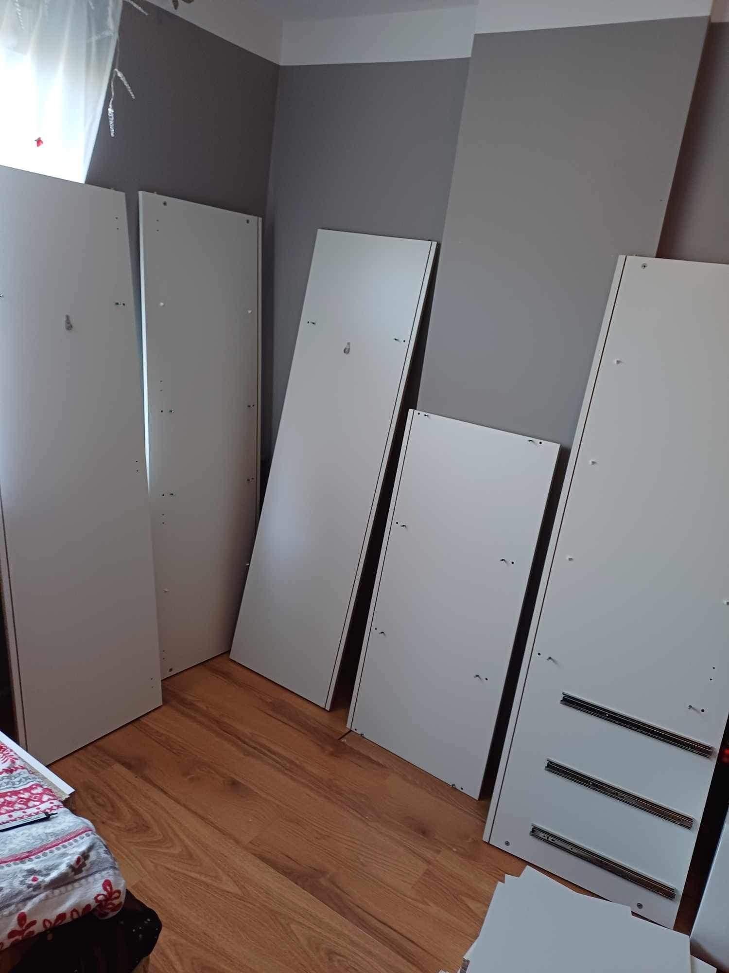 Płyty meblowe w kolorze białym, przygotowane do montażu w Skierniewicach, oparte o szarą ścianę na drewnianej podłodze. Widoczne prowadnice do szuflad i otwory montażowe.