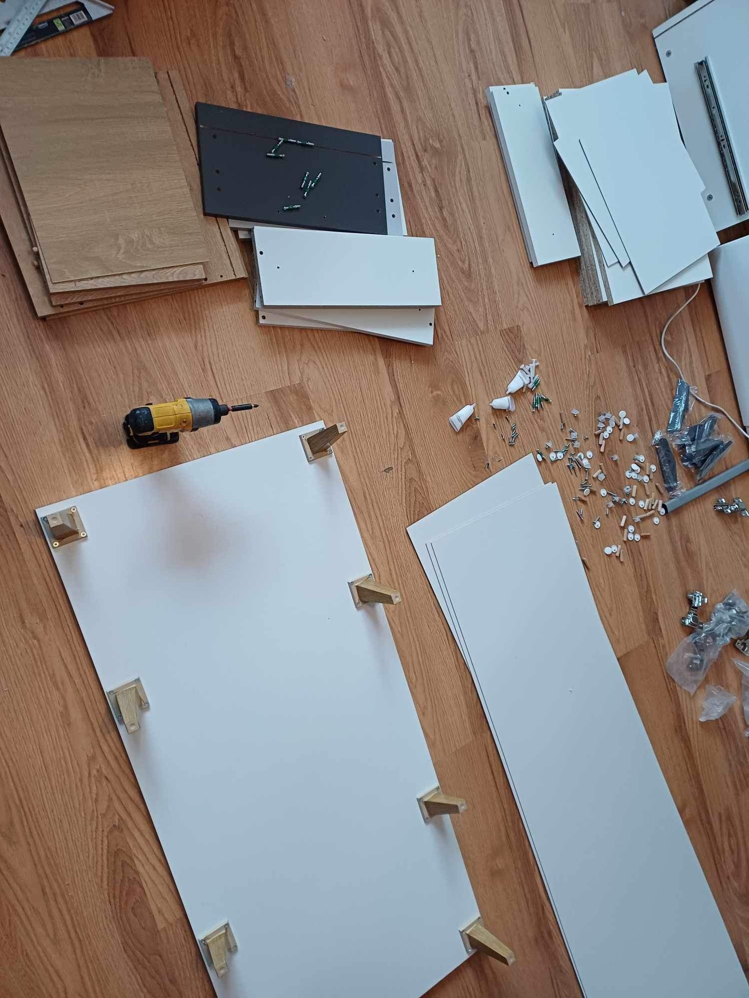 Rozłożone elementy mebli na podłodze z paneli, wiertarka akumulatorowa leży obok białego panelu z zamontowanymi drewnianymi kołkami, widoczne śruby, kołki i inne akcesoria montażowe.