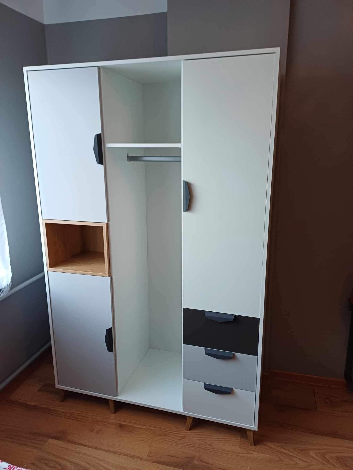 Nowoczesna, biało-szara szafa z drewnianymi akcentami, widoczna na tle dwukolorowej ściany i drewnianej podłogi. Szafa posiada otwartą półkę z jasnego drewna i trzy szuflady w odcieniach szarości.