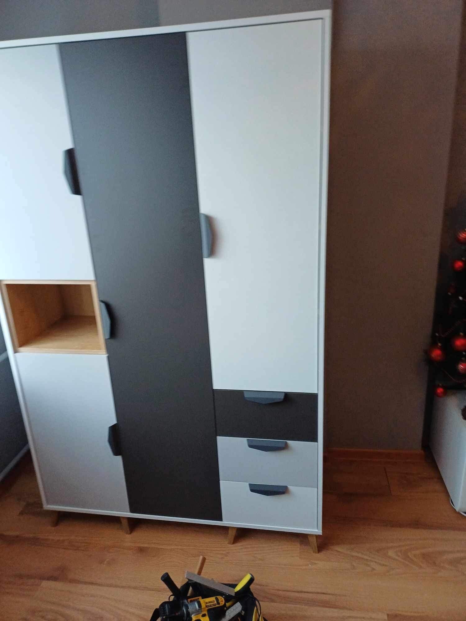 Trójkolorowa szafa z drewnianymi nóżkami, częściowo biała, częściowo szara, z widocznymi narzędziami na podłodze, sugerującymi montaż lub remont.