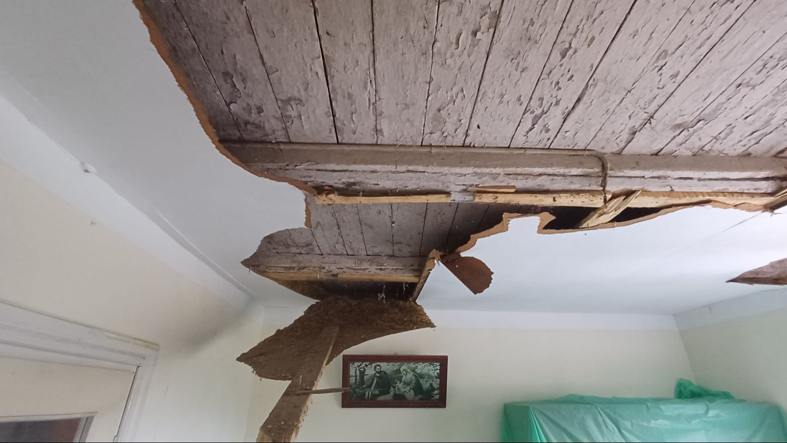 Zdjęcie przedstawia uszkodzony sufit w pokoju, z widocznymi drewnianymi belkami i odpadającym tynkiem. Na ścianie wisi obraz w ramie, a pod ścianą znajduje się mebel przykryty folią ochronną.