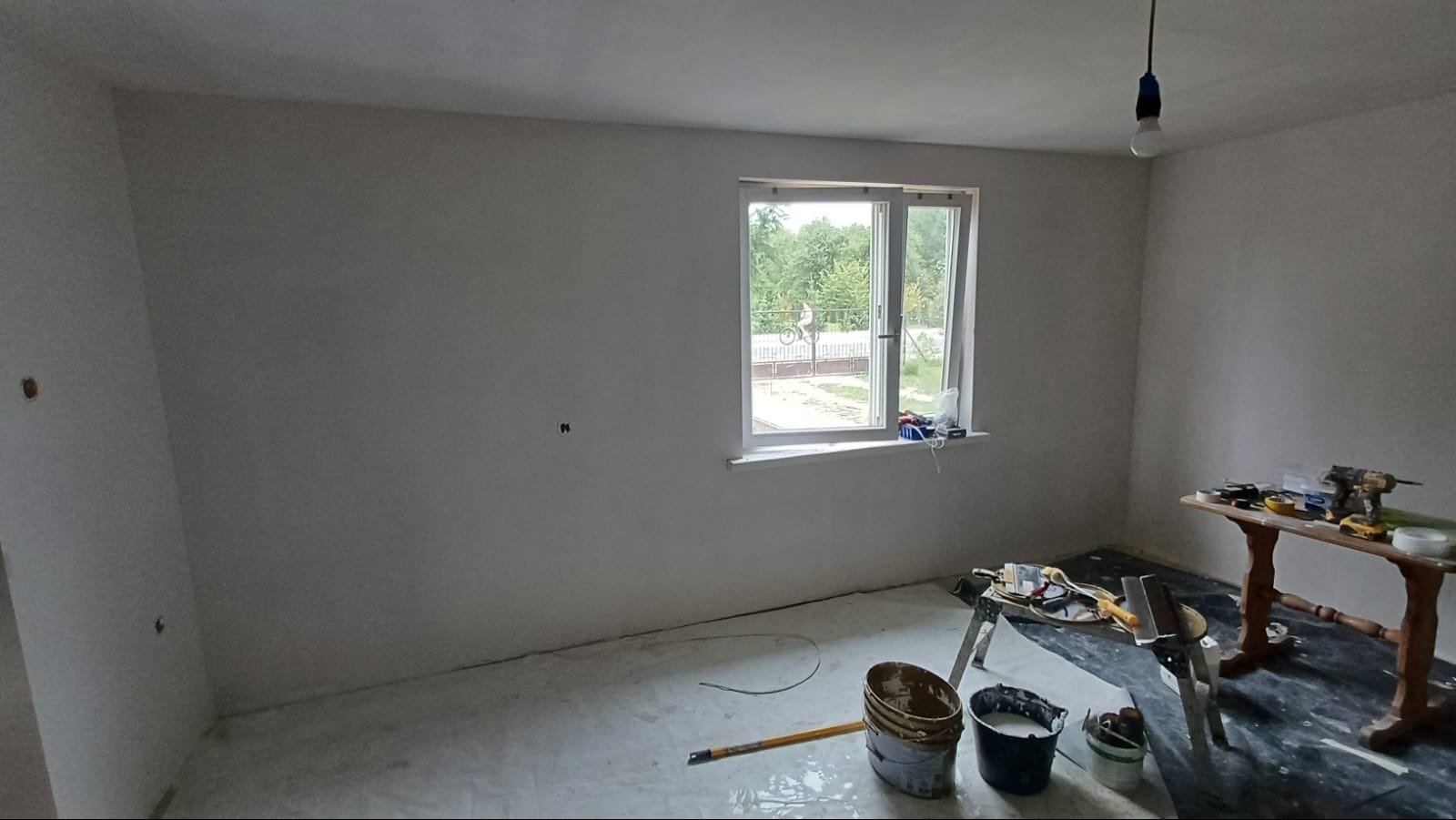 Pokój w trakcie prac wykończeniowych, ściany po nałożeniu gładzi, widoczne narzędzia i wiaderka z masą szpachlową oraz stół z elektronarzędziami, przez okno widok na zieleń.