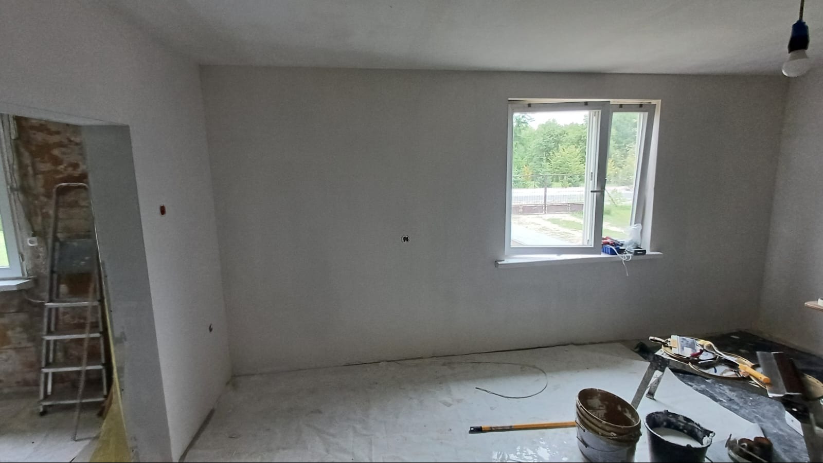 Wnętrze pokoju w trakcie prac wykończeniowych, ściany pokryte świeżą gładzią, widoczne narzędzia malarskie, drabina oraz fragment odsłoniętej ściany z cegły w sąsiednim pomieszczeniu.