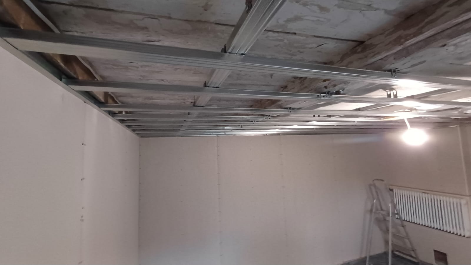 Montaż metalowego stelaża sufitowego pod płyty gipsowo-kartonowe w pomieszczeniu z grzejnikiem i drabiną, widoczne elementy mocujące do sufitu.