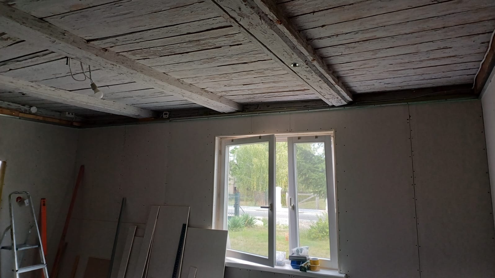 Pokój w trakcie remontu: ściany wyłożone płytami kartonowo-gipsowymi, sufit z drewnianymi belkami pokrytymi łuszczącą się białą farbą, okno z widokiem na zieleń, drabina i materiały budowlane...