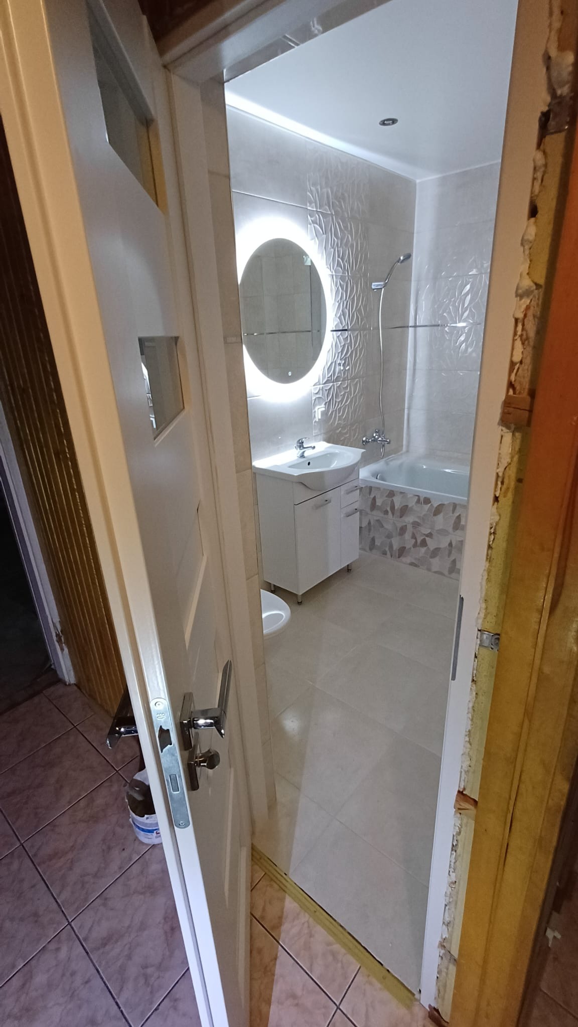 Wykończona łazienka z okrągłym lustrem podświetlanym LED, widok przez otwarte drzwi z widoczną ościeżnicą w trakcie montażu.