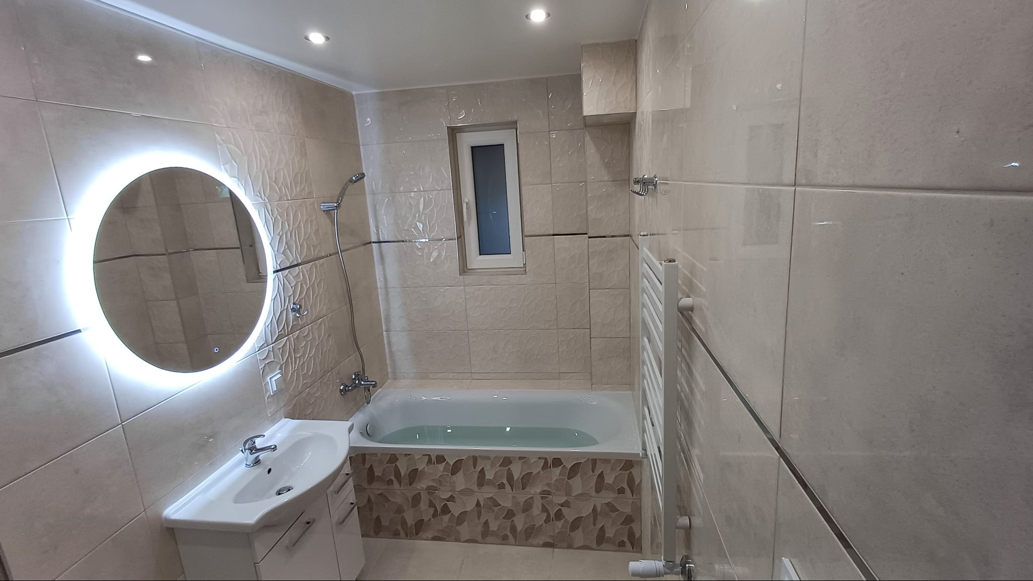 Wykończona łazienka z beżowymi płytkami, okrągłym lustrem z podświetleniem LED, białą umywalką z szafką i wanną z dekoracyjną obudową. W tle widoczny grzejnik drabinkowy i małe okno.