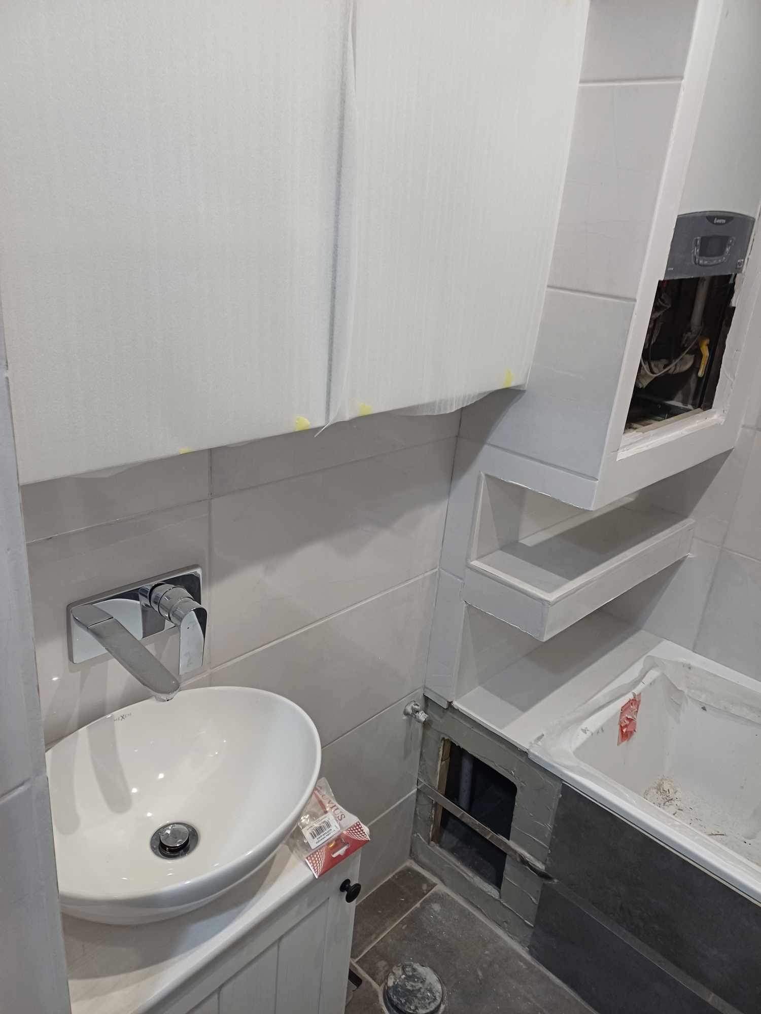 Remont łazienki w trakcie: zabudowa wanny z szarą mozaiką, umywalka nablatowa z baterią, zabudowa szafek zabezpieczona folią malarską, półki w zabudowie z widoczną instalacją.