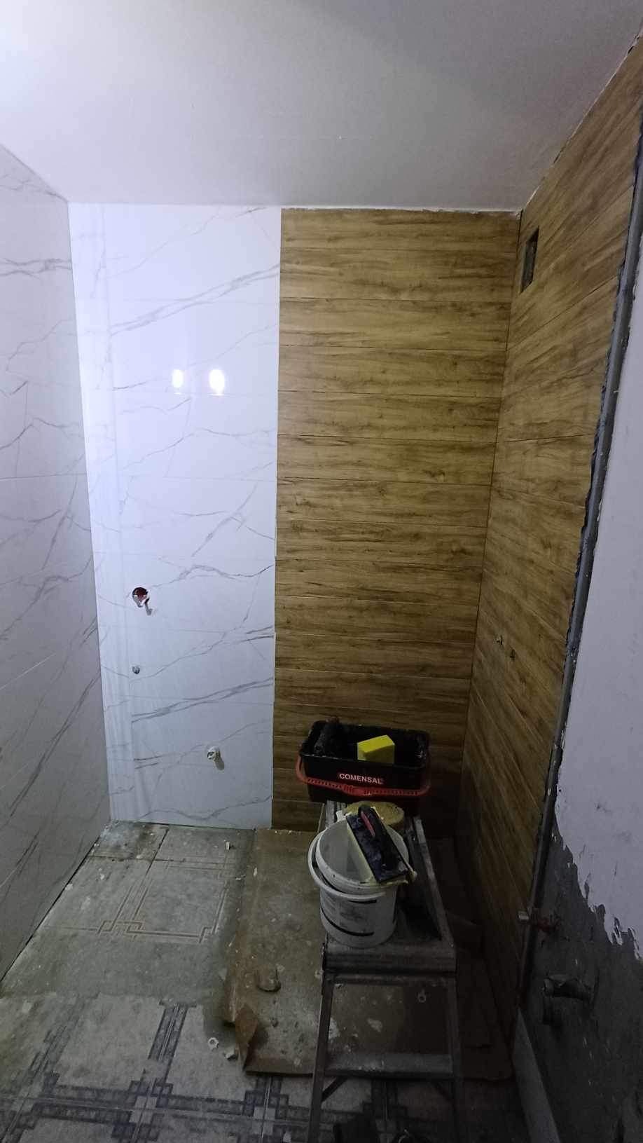 Wnętrze łazienki w trakcie remontu: częściowo wyłożone płytkami imitującymi marmur, częściowo panelami ściennymi o wyglądzie drewna, widoczne narzędzia i wiadro na drabince.