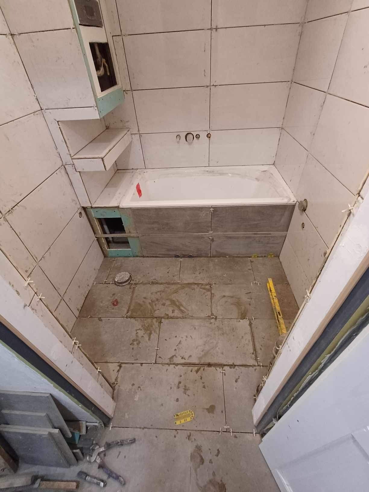 Łazienka w trakcie remontu: układanie płytek ceramicznych na ścianach i podłodze, widoczna wanna i poziomica, białe krzyżyki dystansowe między płytkami.