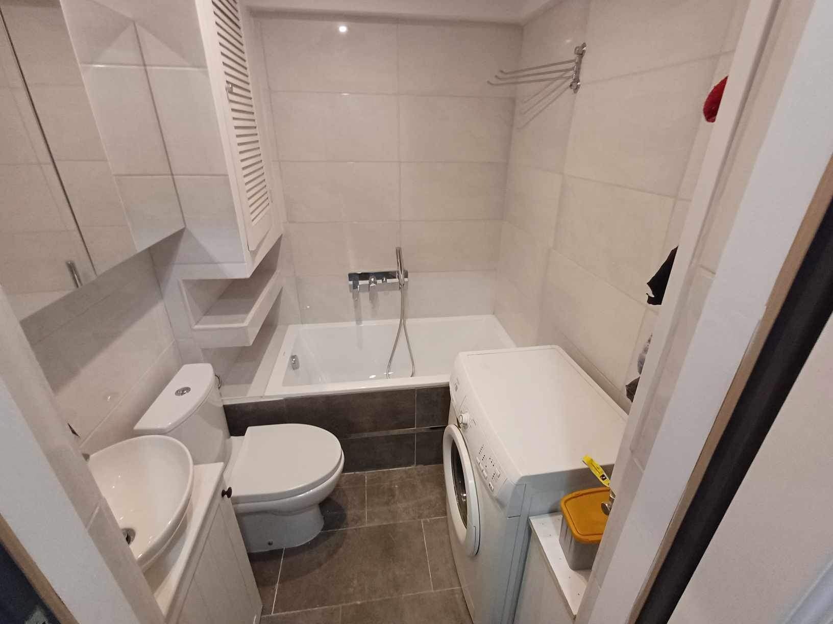 Wykończona łazienka w jasnych płytkach z wanną, umywalką, toaletą i pralką, widok z perspektywy drzwi, z akcentem na minimalistyczne szafki i armaturę.