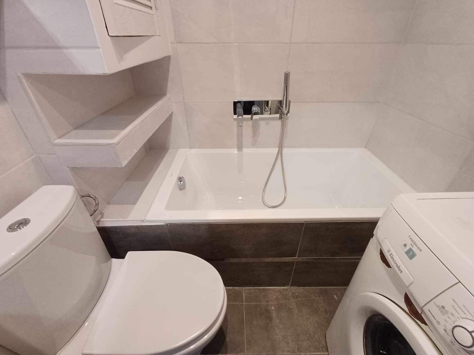 Wykończona łazienka z białą wanną, toaletą, zabudowanymi półkami i pralką Electrolux, płytki w odcieniach bieli i szarości.
