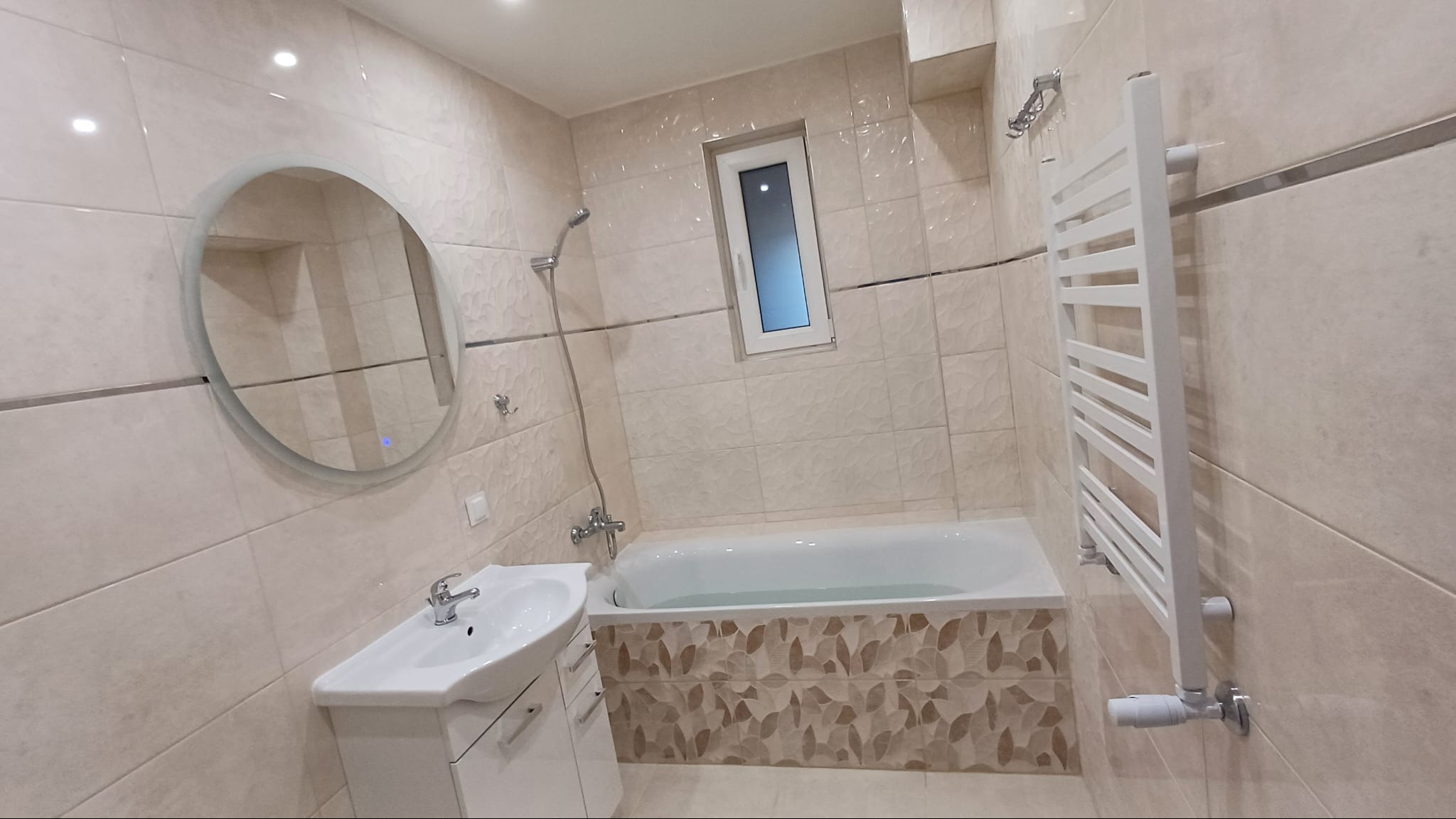 Wykończona łazienka w beżowych płytkach z dekoracyjnym pasem na wannie, okrągłe podświetlane lustro, biała umywalka z szafką i grzejnik drabinkowy.