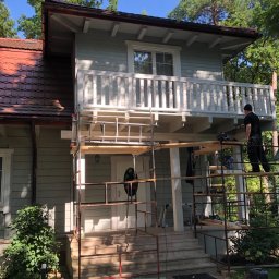 PT. LTD CONSTRUCTION - Malowanie elewacji domu jednorodzinnego z drewnianą konstrukcją balkonu, widoczne rusztowanie i pracownik w trakcie malowania detali balustrady.