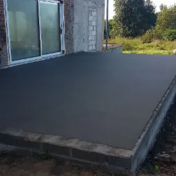 Wylany, świeży betonowy taras przylegający do budynku w stanie surowym otwartym, widoczne bloczki betonowe i częściowo obłożona cegłą ściana z oknem przesuwnym.