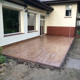 PT. LTD CONSTRUCTION - Wyłożony płytkami imitującymi drewno taras przy domu z beżową elewacją i ceglanym fragmentem ściany. Na tarasie widoczny mop.