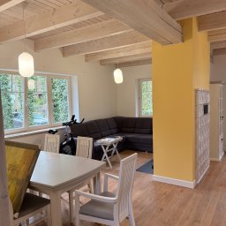 PT. LTD CONSTRUCTION - Jasne wnętrze z drewnianą podłogą i sufitem belkowym. Żółty filar z piecem kaflowym, szary narożnik i białe meble tworzą przytulną przestrzeń.