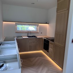 PT. LTD CONSTRUCTION - Montaż kuchni z drewnianymi szafkami i oświetleniem LED. Widoczny fragment podłogi ułożonej w jodełkę i niedokończone szafki po lewej stronie.