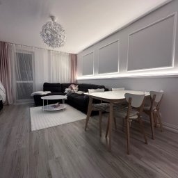 PT. LTD CONSTRUCTION - Jasne wnętrze salonu z drewnianą podłogą, białym dywanem, sofą i stołem z krzesłami. Dekoracyjne listwy ścienne z oświetleniem LED. Nowoczesny żyrandol.
