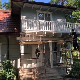 PT. LTD CONSTRUCTION - Dwupiętrowy dom z balkonem w trakcie malowania elewacji, widoczne rusztowania i pracownik w czarnym ubraniu na rusztowaniu.