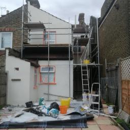 PT. LTD CONSTRUCTION - Malowanie elewacji domu jednorodzinnego z użyciem rusztowania, zabezpieczone okna folią, widoczne wiaderka z farbą i narzędzia malarskie na posesji.