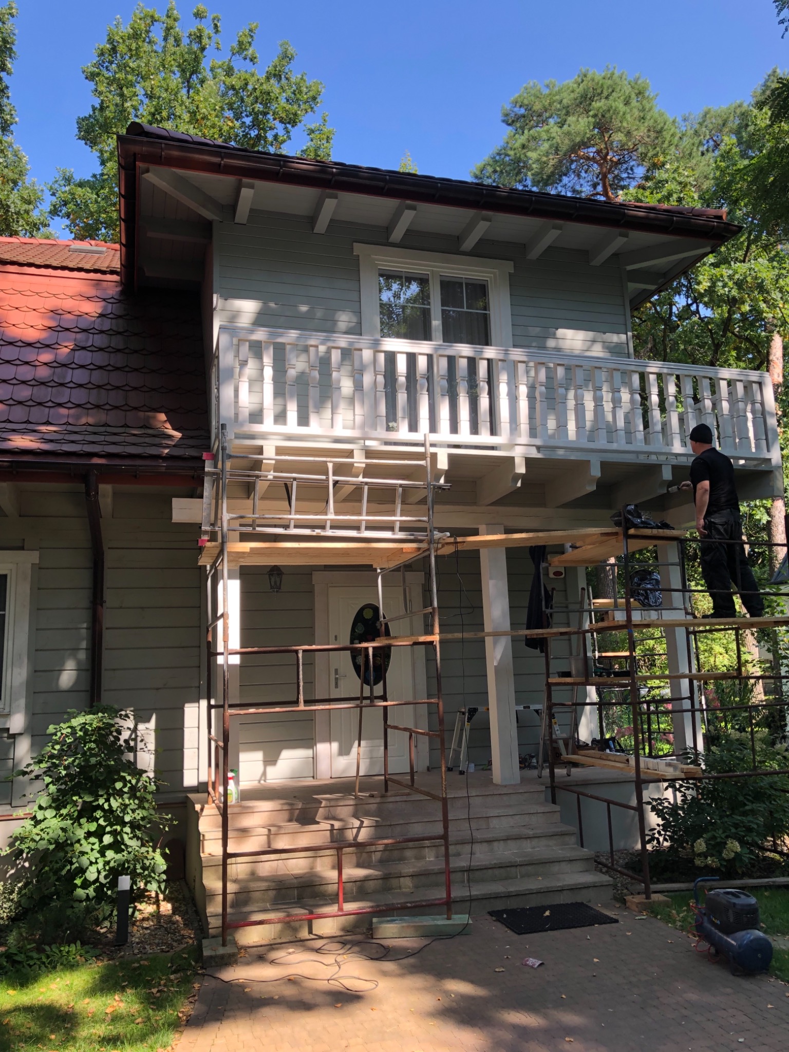 Malowanie elewacji domu jednorodzinnego z drewnianą konstrukcją balkonu, widoczne rusztowanie i pracownik w trakcie malowania detali balustrady.