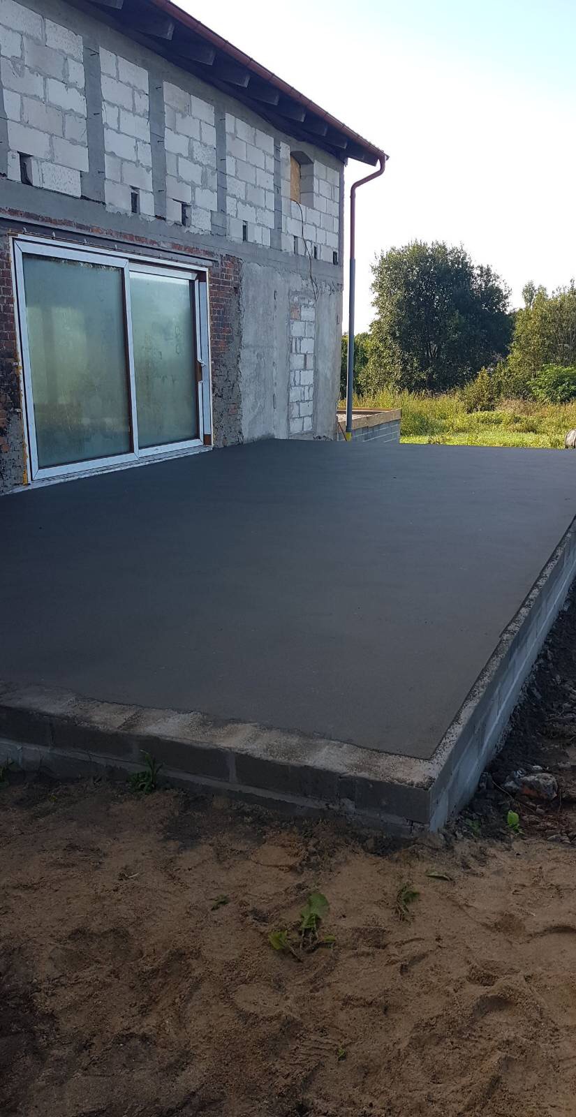 Wylany, świeży betonowy taras przylegający do budynku w stanie surowym otwartym, widoczne bloczki betonowe i częściowo obłożona cegłą ściana z oknem przesuwnym.