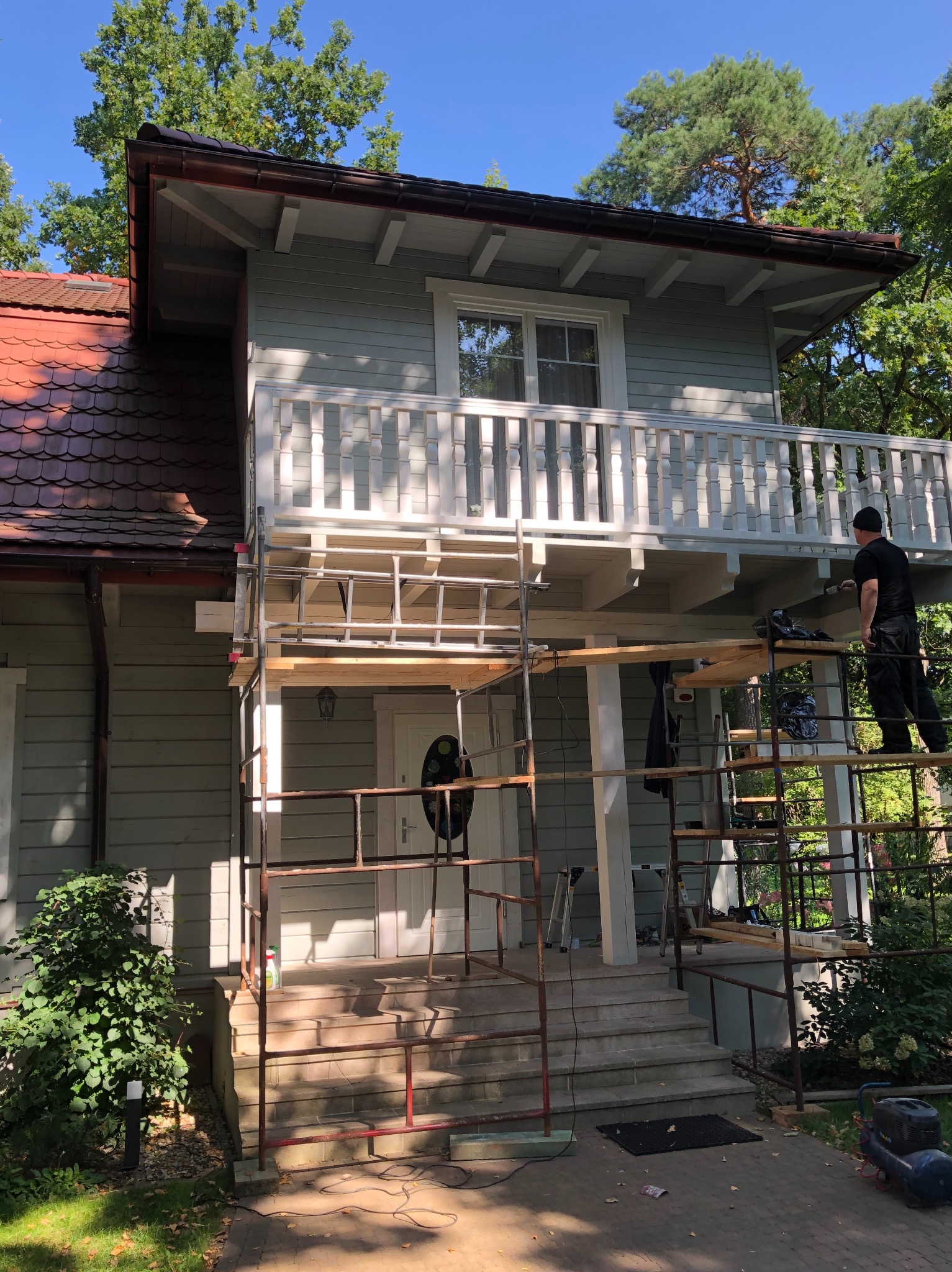 Dwupiętrowy dom z balkonem w trakcie malowania elewacji, widoczne rusztowania i pracownik w czarnym ubraniu na rusztowaniu.