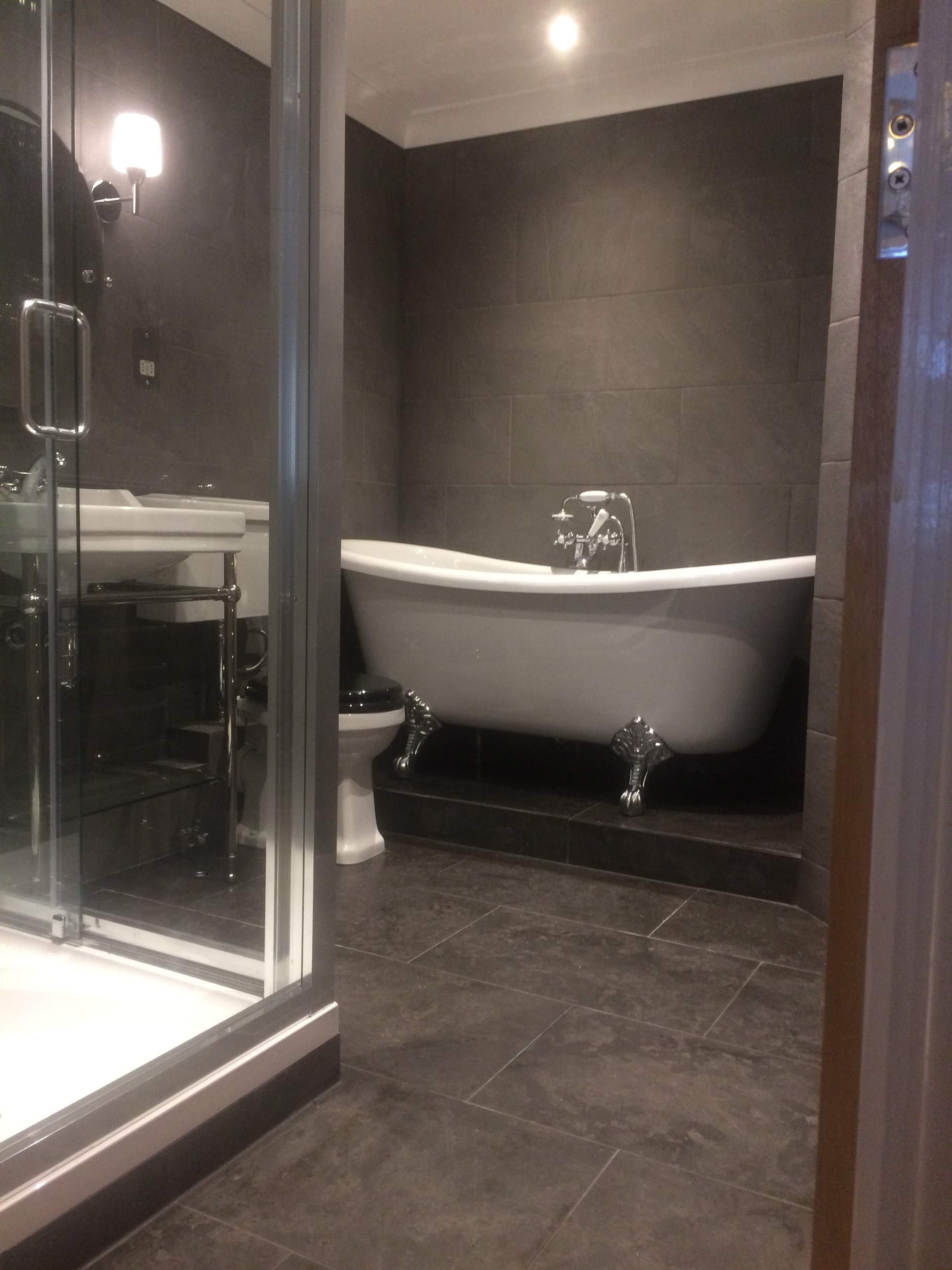 Wykończona łazienka w odcieniach szarości z wolnostojącą wanną na ozdobnych nóżkach, kabiną prysznicową ze szklanymi drzwiami i umywalką na chromowanym stelażu.