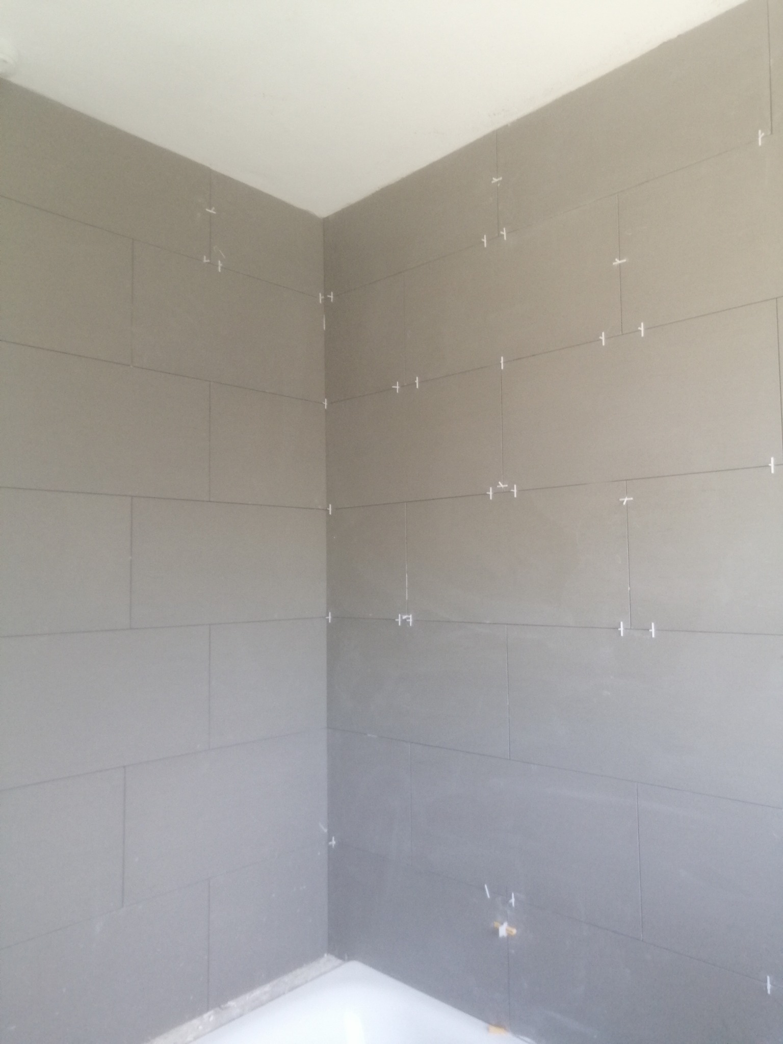 Świeżo ułożone, szare, prostokątne płytki na ścianach w rogu łazienki, z białymi krzyżykami dystansowymi w fugach.