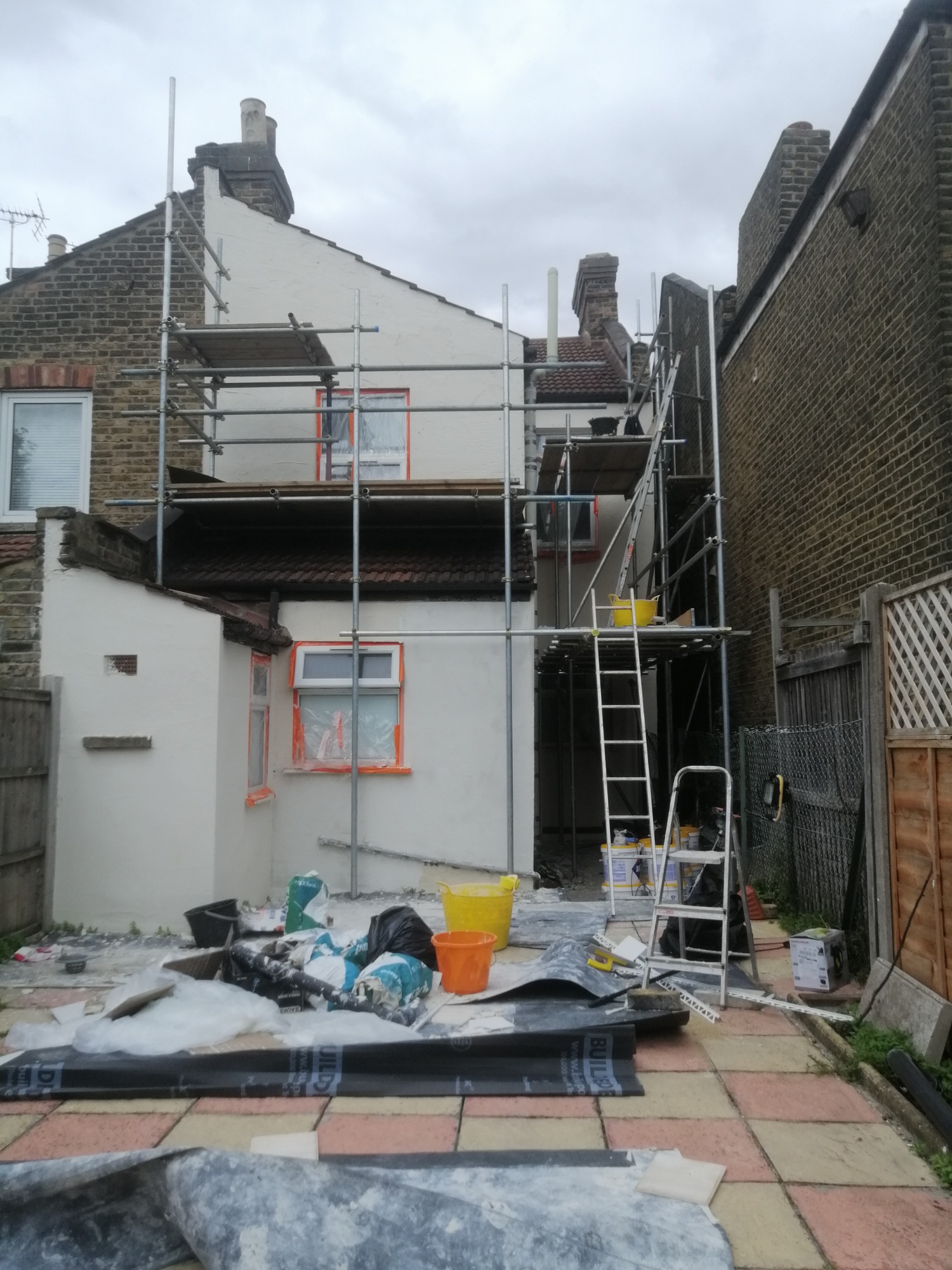 Malowanie elewacji domu jednorodzinnego z użyciem rusztowania, zabezpieczone okna folią, widoczne wiaderka z farbą i narzędzia malarskie na posesji.