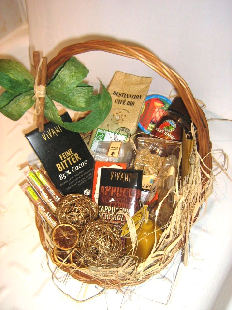 Wiklinowy kosz wypełniony różnorodnymi produktami spożywczymi i dekoracyjnymi, w tym czekoladami Vivani, kawą Destination Cafe Bio, miodem Mount Hagen, kulami dekoracyjnymi, suszonymi pomarańczami...