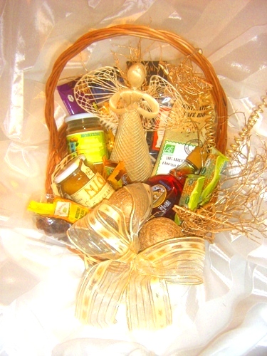 Wiklinowy kosz wypełniony upominkami, w tym słoiczki z miodem, kawa, słodycze i ozdobny aniołek ze słomy, całość przewiązana kremową wstążką w kokardę.