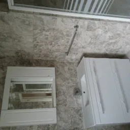 Ukończona łazienka z szarymi marmurkowymi płytkami, białą szafką pod umywalką z trzema drzwiczkami i lustrem z szafką nad umywalką, widoczny fragment kabiny prysznicowej z paskowanym szkłem.