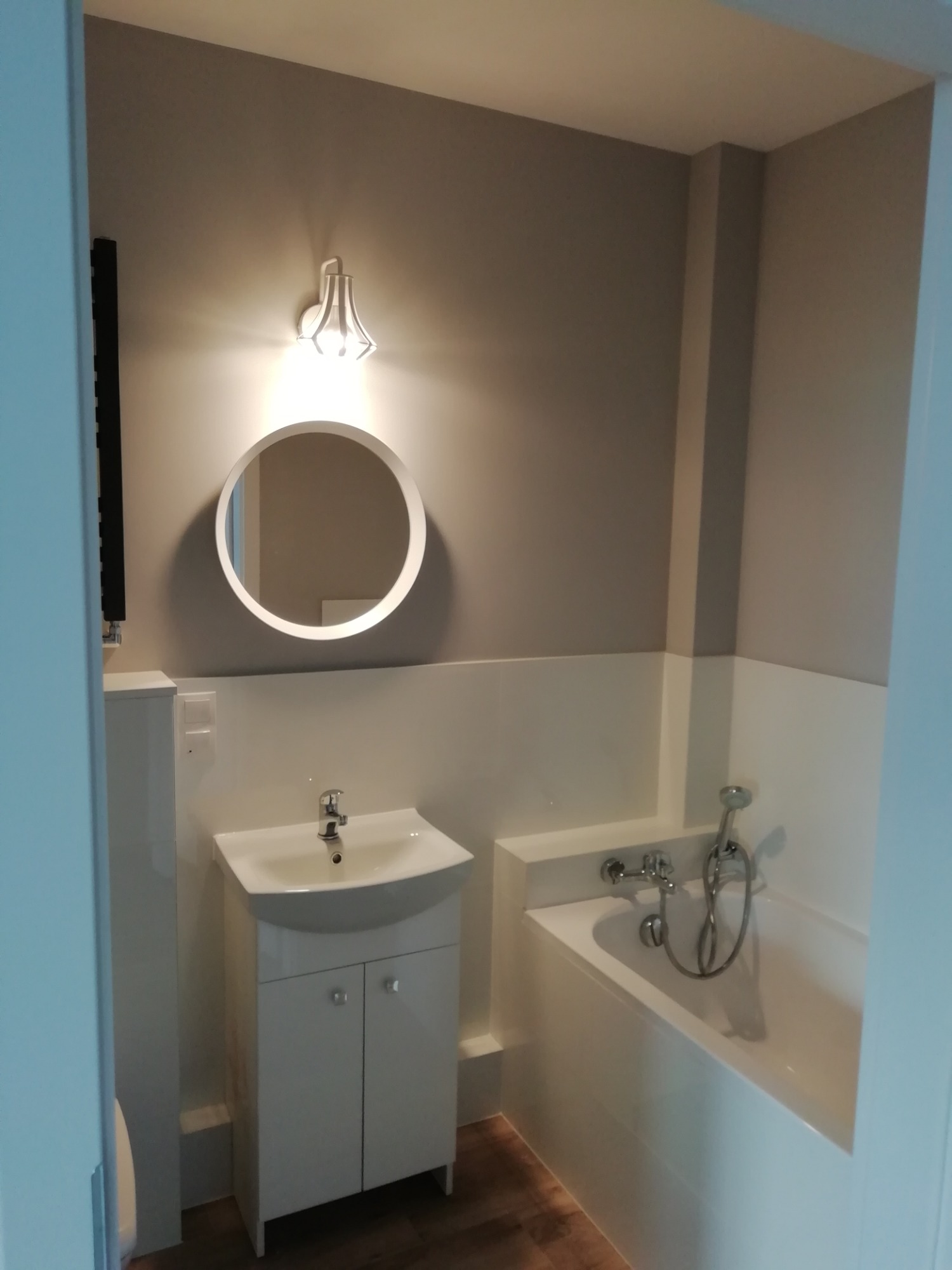 Wykończona łazienka w odcieniach bieli i szarości z okrągłym lustrem, kinkietem nad nim, umywalką z szafką i wanną z armaturą.