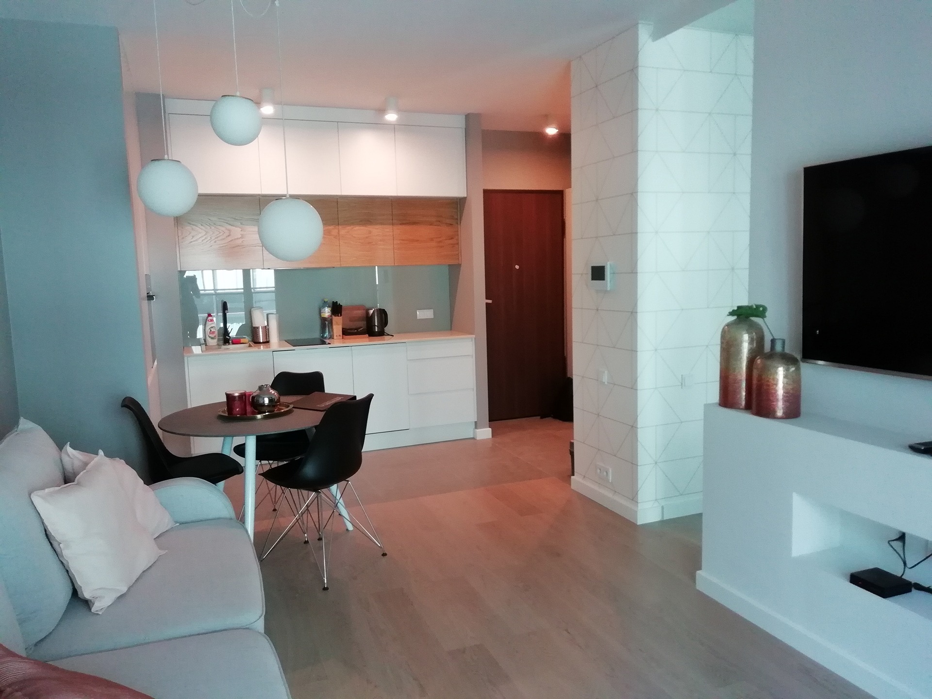 Jasne, nowoczesne wnętrze mieszkania z parkietem, okrągłym stołem i krzesłami Eames, widok na kuchnię z białymi szafkami i drewnianym blatem oraz dekoracyjną kolumną z geometrycznym wzorem.