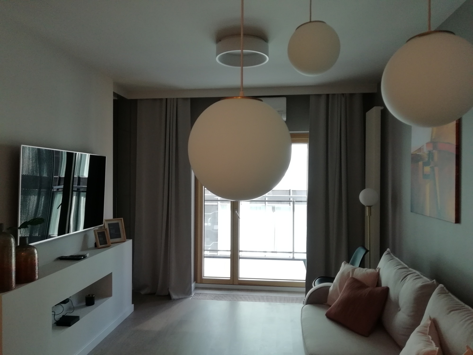 Jasne, przestronne wnętrze salonu z trzema kulistymi lampami wiszącymi, szarymi zasłonami, telewizorem na białej szafce RTV i beżową kanapą z poduszkami.