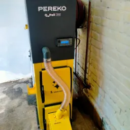 Żółto-czarny piec na pellet marki PEREKO Qm Pell 8kW z podajnikiem, widoczny na tle białej, częściowo odnowionej ściany z cegły i rur instalacyjnych.