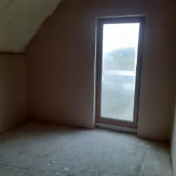Pokój na poddaszu po nałożeniu gładzi, widoczne nierówności na suficie i podłodze, okno z widokiem na zewnątrz.