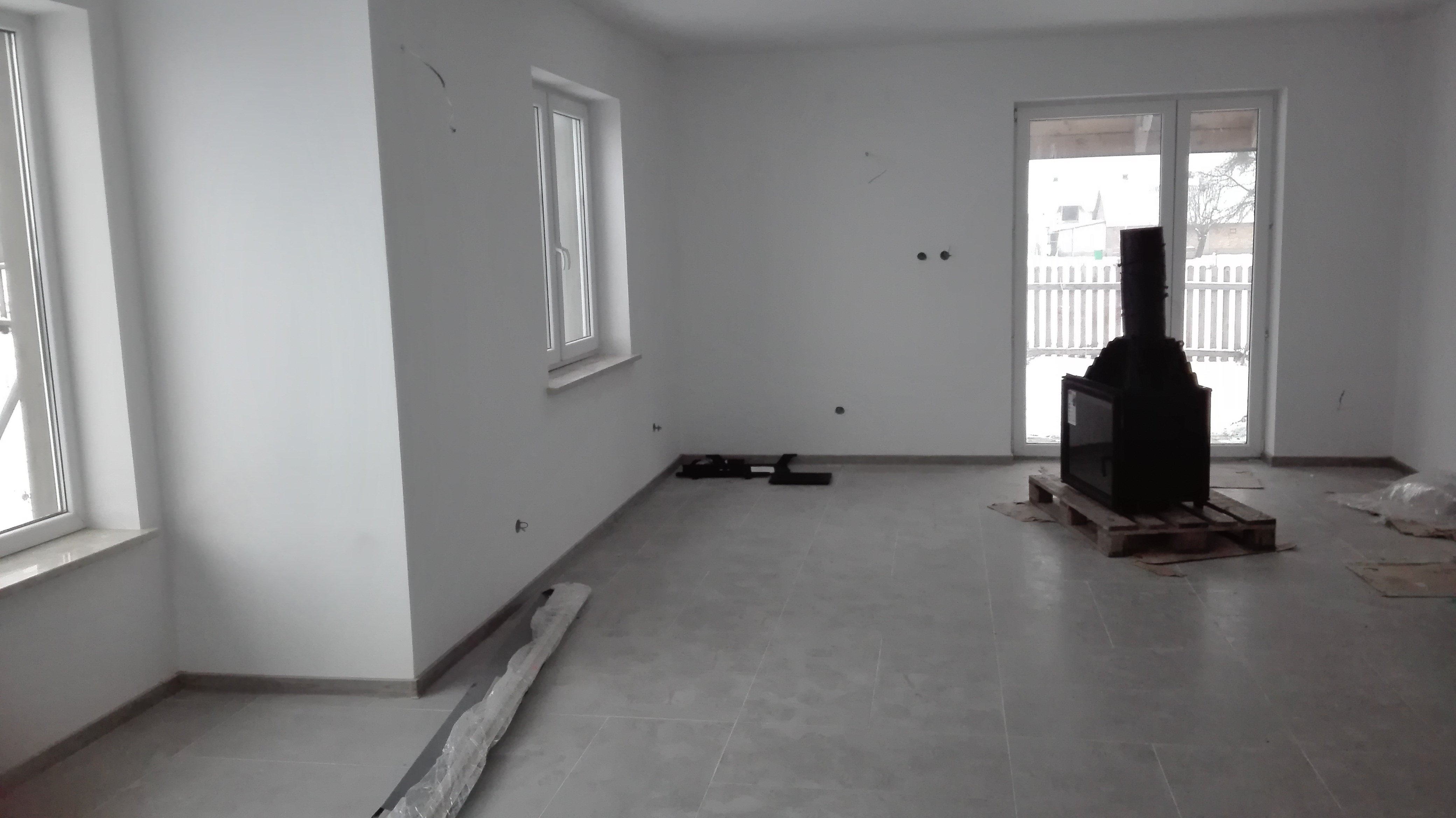 Wnętrze nowo wykończonego pokoju z jasnymi ścianami, szarą podłogą z płytek i zainstalowanym czarnym kominkiem na palecie, widoczne przygotowania do montażu.