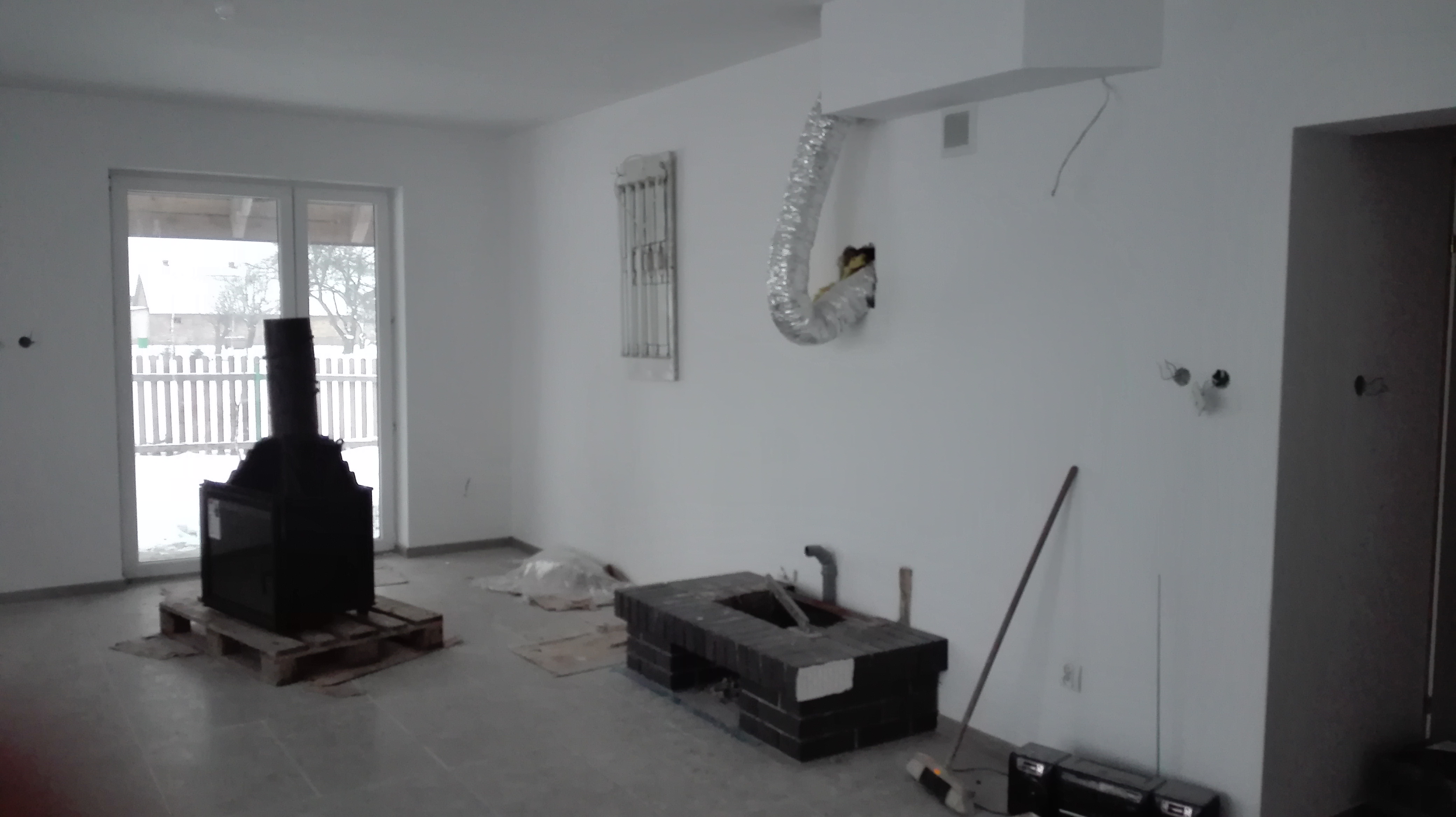 Wnętrze w trakcie prac wykończeniowych: czarny piec kominkowy na palecie, murowana podstawa kominka z czarnej cegły, wentylacja w suficie, surowe ściany i podłoga.