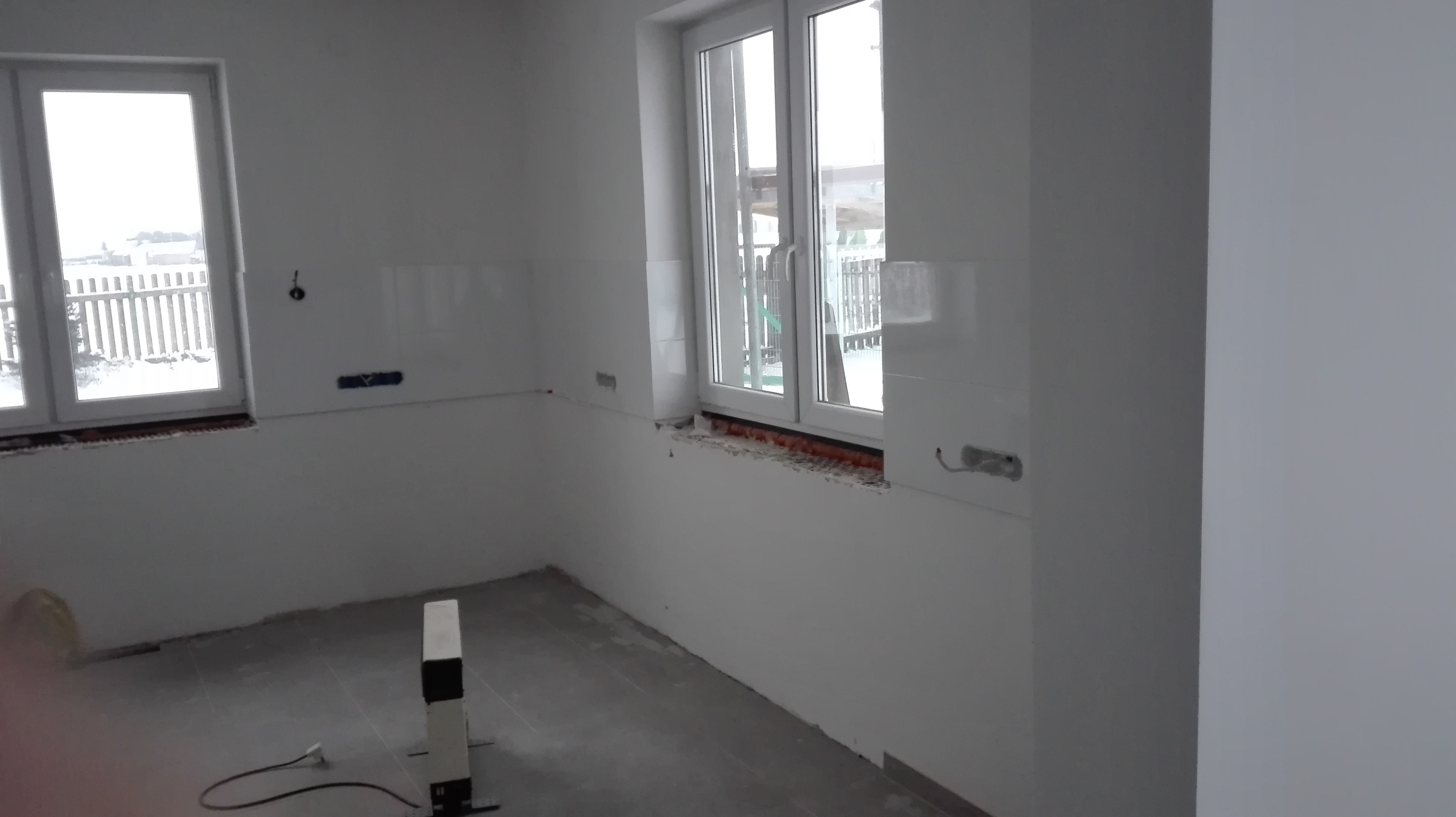 Wnętrze w trakcie remontu, widoczne białe ściany z fragmentami płytek, okna z widokiem na zimowy krajobraz i grzejnik elektryczny na podłodze.