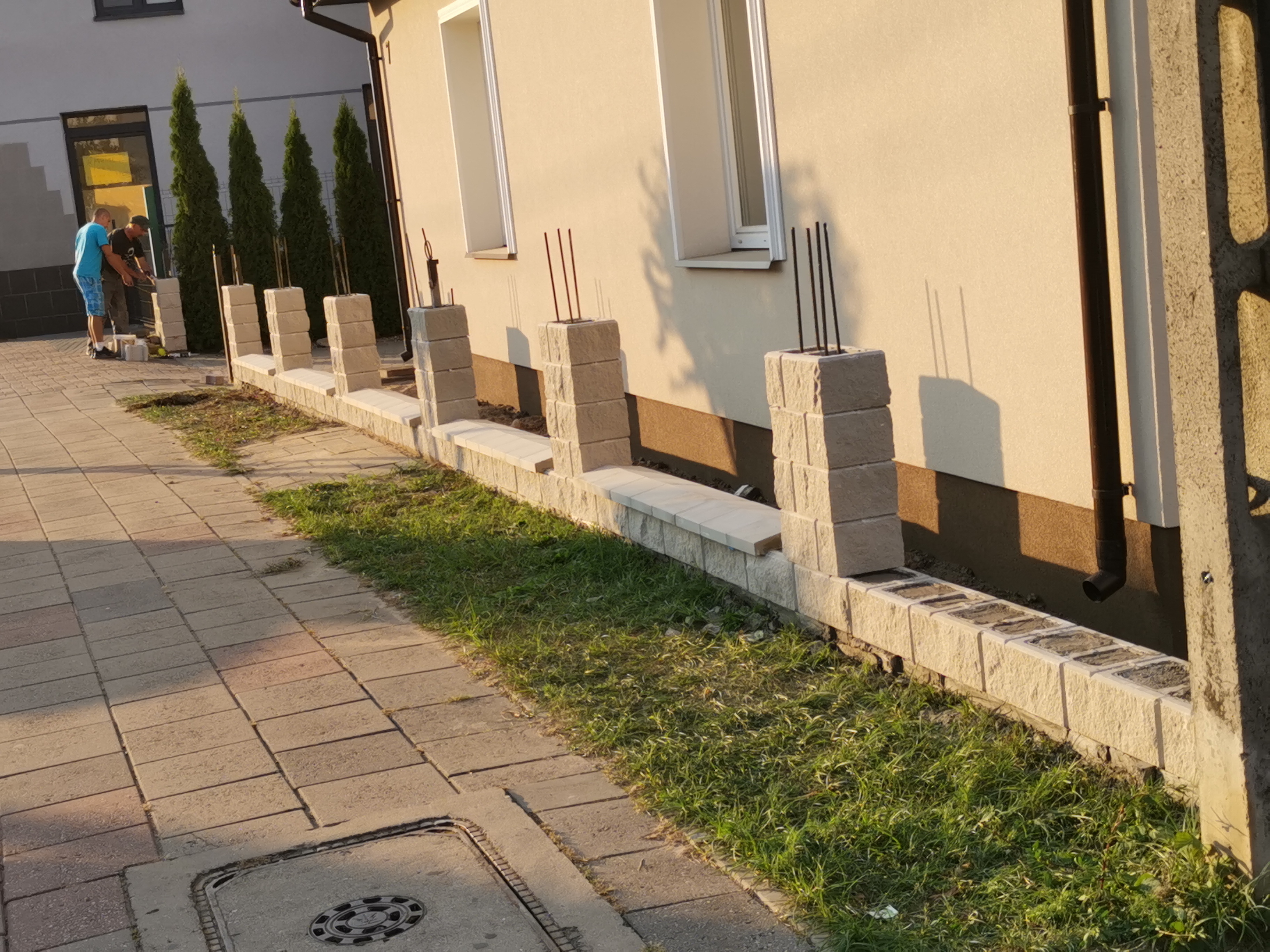 Budowa ogrodzenia z bloczków betonowych przy budynku z widocznymi prętami zbrojeniowymi w słupkach, dwaj pracownicy w tle.