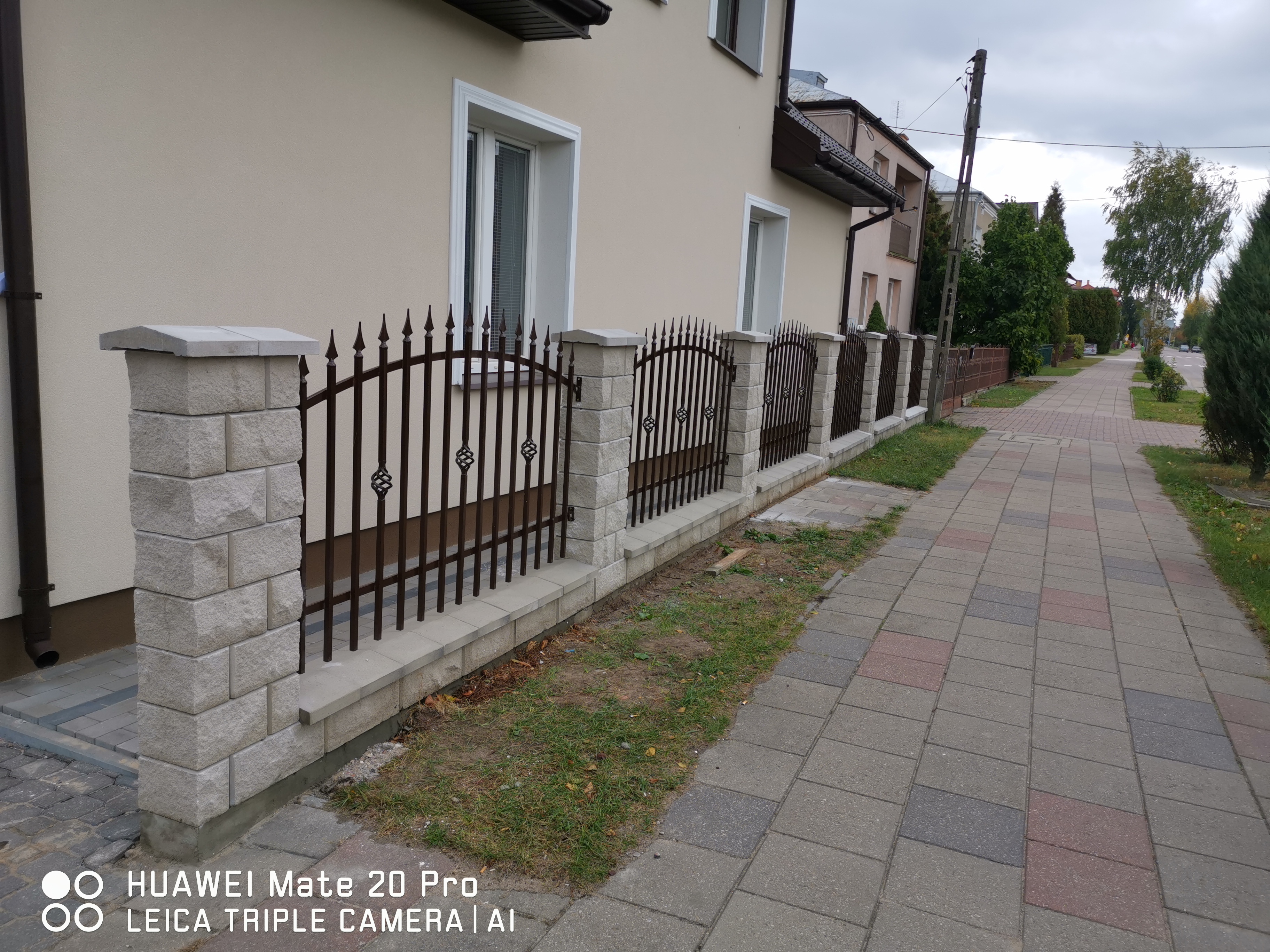 Nowoczesne ogrodzenie z metalowymi przęsłami i kamiennymi słupkami, widoczne z perspektywy chodnika, z domem w tle.