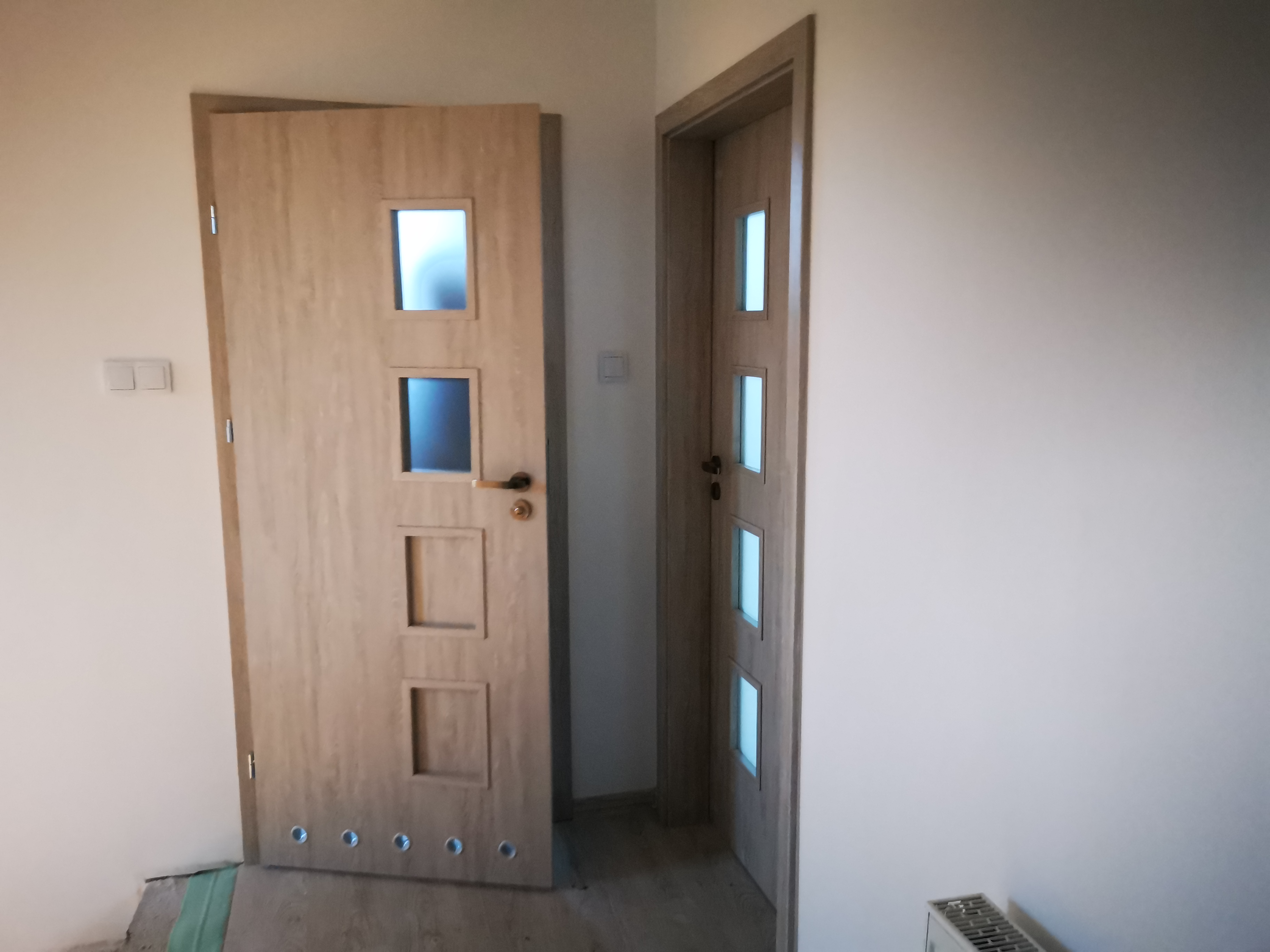 Dwie drewniane drzwi wewnętrzne z przeszkleniami, jedna z kwadratowymi szybkami, druga z podłużnymi, zamontowane w jasnym pomieszczeniu, widoczne ślady prac wykończeniowych na podłodze.