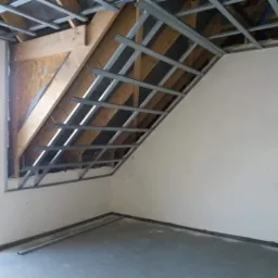 Montaż metalowej konstrukcji pod płytę gipsowo-kartonową na poddaszu, widoczne drewniane elementy konstrukcyjne dachu i surowy stan ścian, przygotowanie do dalszych prac wykończeniowych.